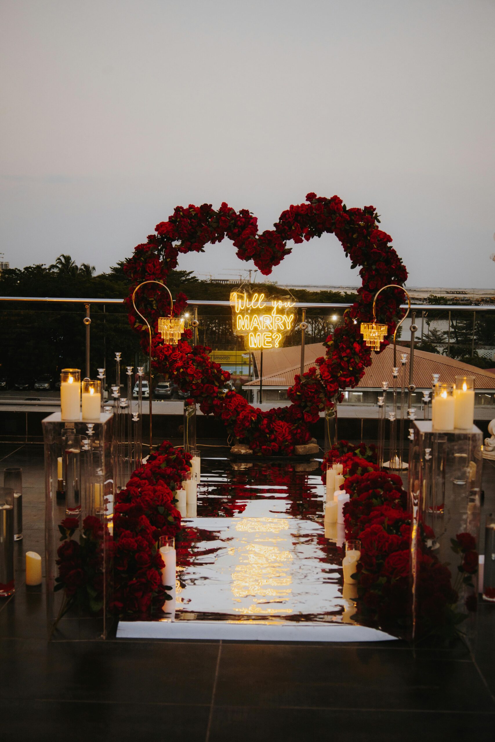 Romantic rooftop proposal setup at sunset with red roses, mirrored aisle, candles, and a “Will You Marry Me?” neon sign.